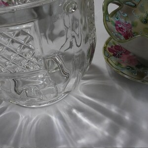 May include: A clear glass bowl with a decorative pattern, sitting on a white surface. The bowl has a wide, round shape and a textured surface. The bowl is reflecting light, creating a shimmering effect.