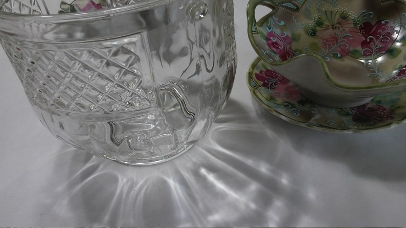 May include: A clear glass bowl with a decorative pattern, sitting on a white surface. The bowl has a wide, round shape and a textured surface. The bowl is reflecting light, creating a shimmering effect.
