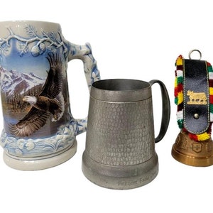 Puede incluir: Tres jarras, una con un diseño de águila azul y blanco, otra con un acabado de peltre y el texto "Freuden-Krug", y la última con un diseño de vaca y una borla colorida.