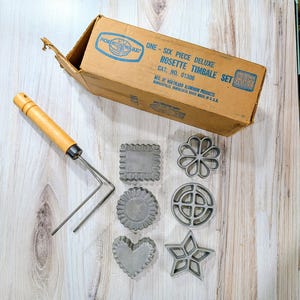 May include: A vintage Nordic Ware Rosette Timbale Set with six decorative cookie molds and a wooden-handled dipping tool. The set includes various shapes like a star, heart, and flower, all in a cardboard box.