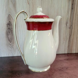 Puede incluir: Tetera de cerámica blanca con tapa roja y dorada y ribete dorado. La tetera tiene un diseño de rayo estilizado.