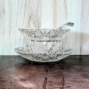 Könnte beinhalten: Ein klares, geschliffenes Glasservice, bestehend aus einer Schüssel, einer kleineren Schüssel, einer Untertasse und einem Löffel. Das Set weist komplizierte geometrische und florale Muster auf. Der Löffel liegt auf dem Rand der Schüssel. Das Set wird auf einer dunklen Oberfläche präsentiert.