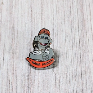 May include: A pin featuring a cartoon monkey wearing an orange hat and holding a banana. The monkey is gray with blue eyes and a red mouth. The banana has the words "NANA PUDDIN" in white.