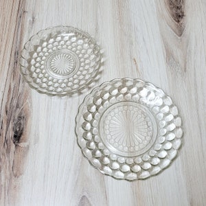 May include: Two clear glass plates with a raised dot pattern. The plates are on a light wood surface.