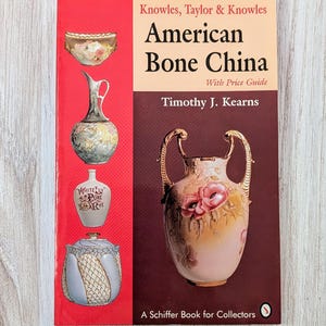 May include: Book cover for "American Bone China" by Timothy J. Kearns. The cover features a red background with four antique porcelain pieces: a bowl, a pitcher, a bottle, and a vase. The book is a Schiffer Book for Collectors.