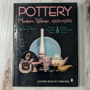 May include: A black book titled "Pottery: Modern Wares 1920-1960" by Leslie Piña. The book cover features a collection of colorful pottery, including a blue and white plate, a striped vase, and a yellow teapot.