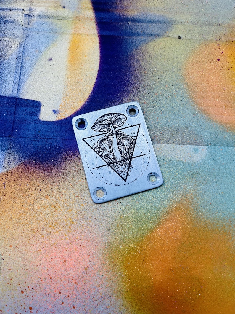 Peut inclure: Plaque m&eacute;tallique argent&eacute;e avec une illustration noire d&eacute;taill&eacute;e de champignons &agrave; l'int&eacute;rieur d'un triangle. La plaque comporte quatre trous, un dans chaque coin. Le fond est un motif peint &agrave; la bombe color&eacute; avec des teintes bleues, oranges et jaunes.