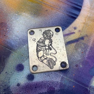 Peut inclure: Plaque m&eacute;tallique argent&eacute;e rectangulaire avec quatre trous, orn&eacute;e d'une illustration noire d&eacute;taill&eacute;e d'un scaphandre vintage. Le plongeur est repr&eacute;sent&eacute; flottant, avec des bulles et un tuyau. La plaque a un aspect us&eacute; et textur&eacute;.
