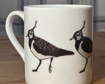 Può includere: Una tazza in ceramica bianca con un'illustrazione in bianco e nero di due uccelli. Gli uccelli sono rivolti in direzioni opposte e hanno gambe lunghe e sottili.