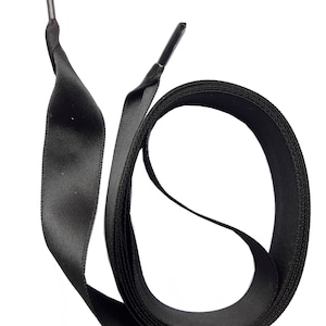 Może przedstawiać: Para czarnych sznurowadeł z metalowymi agletami. Sznurówki wykonane są z płaskiego, tkanego materiału i zwinięte w pętlę. Aglety są w ciemnoszarym kolorze.