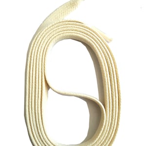 Puede incluir: Cordón de zapato plano de color crema, enrollado en un bucle, sobre un fondo blanco. El cordón tiene una textura tejida y una apariencia firme y duradera. La puntera es visible al final del cordón.