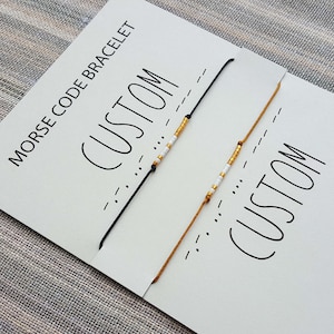 Op de afbeelding: Twee gepersonaliseerde Morsecode-armbanden op witte kaartjes. De armbanden zijn gemaakt met witte en gouden kralen op zwart en bruin touw. De kaartjes zeggen "Morsecode-armband" en "Aangepast".
