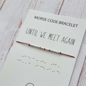 May include: A red string bracelet with silver beads that spells out "Until We Meet Again" in Morse code. The bracelet is on a white card with the phrase "Morse Code Bracelet" and the message "Until We Meet Again" printed on it.