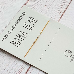 Puede incluir: Una tarjeta blanca con el texto "Morse Code Bracelet Mama Bear" y una pulsera de cuerda dorada con cuentas blancas. La pulsera está anudada y los extremos cuelgan. La tarjeta tiene un logotipo en blanco y negro en la esquina inferior derecha.