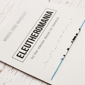 Puede incluir: Un brazalete de cuerda azul y blanco con cuentas negras. El brazalete está sobre una tarjeta blanca con el texto "MORSE CODE BRACELET ELEUTHEROMANIA (n) the intense desire for freedom."