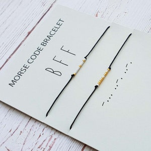 以下が含まれることがあります： 白と金のビーズで「BFF」をモールス信号で綴った、黒い紐のブレスレット2本。ブレスレットは、「モールス信号ブレスレット」と黒で書かれた白いカードに飾られています。