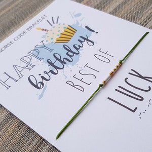 Puede incluir: Un brazalete de cuerda verde con cuentas blancas y doradas que deletrean "BEST OF LUCK" en código morse. El brazalete está sobre una tarjeta blanca con el texto "HAPPY BIRTHDAY!" y una ilustración de una magdalena.