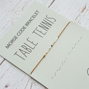 Op de afbeelding: Een wit en gouden kralen armband met een bruin koord. De armband ligt op een kaart met de tekst "Morse Code Armband Tafeltennis".