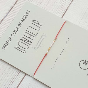 Puede incluir: Pulsera de cuerda roja con cuentas blancas y doradas que deletrean "BONHEUR" en código morse. La tarjeta dice "Pulsera código morse BONHEUR felicidad".