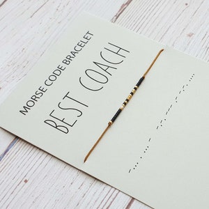 Puede incluir: Un brazalete de cuentas doradas y negras con el mensaje "BEST COACH" escrito en código Morse en una tarjeta blanca. La tarjeta también incluye la frase "MORSE CODE BRACELET".