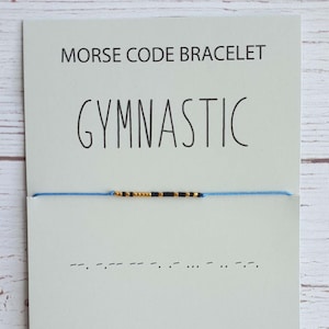 May include: A blue and gold beaded bracelet with black beads spelling out the word "GYMNASTIC" in Morse code. The bracelet is displayed on a white card with the words "MORSE CODE BRACELET" and the phrase "GYMNASTIC" written in black.