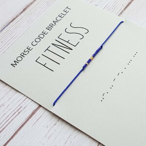 May include: A blue string bracelet with gold beads spelling out "FITNESS" in Morse code. The bracelet is on a white card with the text "MORSE CODE BRACELET" and the word "FITNESS" in black.