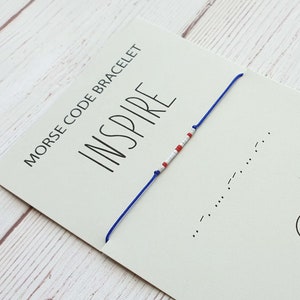 May include: A blue and white beaded bracelet with the word "INSPIRE" written on a card. The bracelet is made with red, white, and blue beads and is tied with a blue string. The card also includes the text "Morse Code Bracelet".