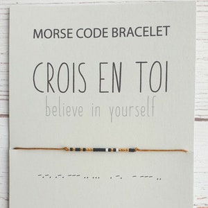 Puede incluir: Un brazalete de cuerda dorada con cuentas negras, blancas y doradas. El brazalete está atado en un nudo y tiene una tarjeta adjunta. La tarjeta dice "Pulsera de código Morse" y "Crois en toi" en francés, que se traduce como "Cree en ti mismo" en inglés.