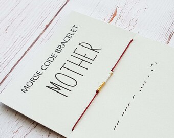 Könnte beinhalten: Ein rotes Armband aus Schnur mit weißen und goldenen Perlen, die "MOTHER" in Morsecode buchstabieren. Das Armband befindet sich auf einer weißen Karte mit den aufgedruckten Worten "Morse Code Bracelet" und "MOTHER".