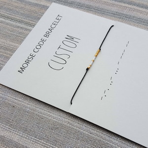 Op de afbeelding: Een witte kaart met zwarte tekst die "Morse Code Armband (Custom)" zegt. Een zwart koordarmband met witte en gouden kralen ligt op de kaart. De kaart heeft ook een klein logo met de letters "WH" in een cirkel.