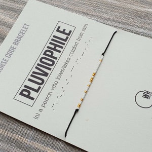 May include: A white and gold beaded bracelet with a black string. The bracelet is on a card that says "Morse Code Bracelet" and "Pluviophile (n) a person who loves/takes comfort from rain."