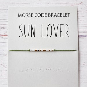 Puede incluir: Un brazalete de cuerda verde con cuentas blancas, doradas y negras. El brazalete deletrea "SUN LOVER" en código Morse. La tarjeta dice "Pulsera de código Morse SUN LOVER".