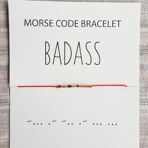 May include: A red string bracelet with gold and black beads spelling out the word "BADASS" in Morse code. The bracelet is displayed on a white card with the word "BADASS" printed in black and the phrase "MORSE CODE BRACELET" printed above it.