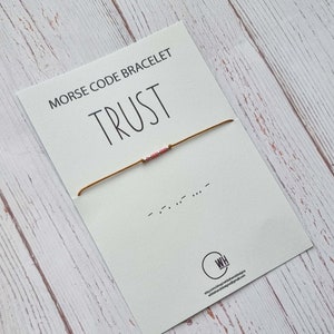 Puede incluir: Un brazalete de cuerda marrón con cuentas rosadas que deletrean la palabra "TRUST" en código Morse. El brazalete se muestra en una tarjeta blanca con la palabra "TRUST" impresa en negro y la frase "MORSE CODE BRACELET" impresa en negro.