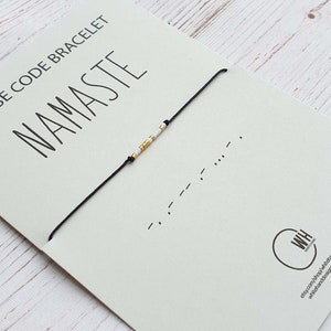 Può includere: Un braccialetto di corda nero con perline bianche e dorate che compongono la parola "namaste" in codice Morse. Il braccialetto è su una carta bianca con le parole "Morse Code Bracelet" e "namaste" stampate su di essa.