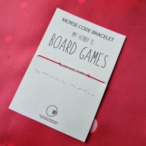 May include: A white card with the text "Morse Code Bracelet" and "My hobby is Board Games". A red string bracelet with silver and pink beads is attached to the card. The bracelet is in Morse code and spells out "Board Games".
