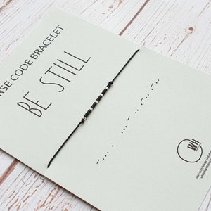 May include: A black string bracelet with white beads spelling out "BE STILL" in Morse code. The bracelet is on a card with the text "Morse Code Bracelet BE STILL".
