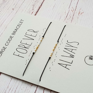 Pode incluir: Duas pulseiras pretas com código Morse, apresentadas num cartão branco. O cartão diz "FOREVER ALWAYS" e "Morse Code Bracelet". As pulseiras têm contas douradas e brancas.