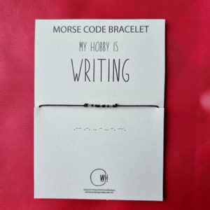 Può includere: Un biglietto bianco con testo nero che recita "Braccialetto in codice Morse" e "Il mio hobby è SCRIVERE". Un braccialetto di corda nero con perline argentate è mostrato sul biglietto. Il braccialetto è in codice Morse e scrive la parola "SCRIVERE".