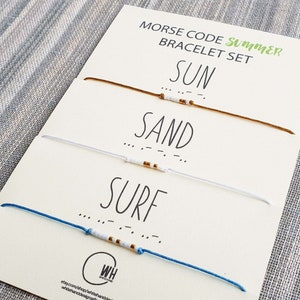 Puede incluir: Un conjunto de tres pulseras de amistad con las palabras "SUN", "SAND" y "SURF" escritas en código Morse usando cuentas blancas y doradas en cordones marrones, blancos y azules.