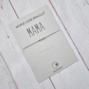 Może przedstawiać: Fioletowa bransoletka ze sznurka z srebrnymi koralikami, które tworzą napis "MAMA" w alfabecie Morse'a. Bransoletka znajduje się na białej kartce z nadrukiem "Bransoletka w alfabecie Morse'a" i "MAMA".