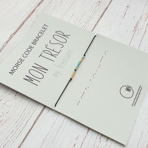 Puede incluir: Una tarjeta blanca con texto negro que dice "Morse Code Bracelet - Mon Trésor - my treasure". Una pulsera de cuerda negra con cuentas turquesa y doradas está unida a la tarjeta.