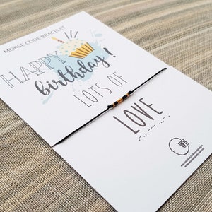 Puede incluir: Una tarjeta blanca con el texto "Pulsera de código Morse" y "¡Feliz cumpleaños! Mucho amor" en letras negras. Una pulsera de cuerda negra con cuentas amarillas y negras está unida a la tarjeta.