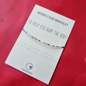 May include: A white card with black text that reads "Morse Code Bracelet" and "I'd help you bury the body". A black and white beaded bracelet is displayed on the card. The bracelet is made of small white beads with black and gold accents.