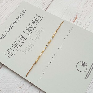 Puede incluir: Un brazalete de cuentas doradas y blancas con el mensaje "HEUREUX ENSEMBLE happy together" en código morse en una tarjeta blanca.
