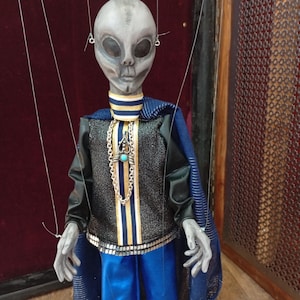 May include: A gray alien puppet with large black eyes, wearing a black jacket, blue pants, and a blue cape. The puppet has a silver chain with a turquoise pendant. The puppet is suspended by strings.