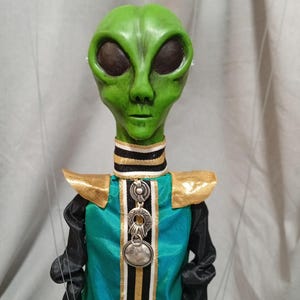 May include: A green alien puppet with large black eyes and a detailed face. The puppet wears a teal and black outfit with gold accents and a silver pendant. The puppet is suspended by strings.