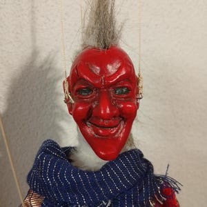 May include: A red puppet with a mohawk, a mischievous grin, and a blue and gold patterned scarf. The puppet has a red face with a white beard and is wearing a red and gold costume.