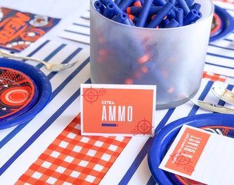 May include: A table setting for a Nerf party with blue and white striped placemats, red and white checkered placemats, blue and white plates, and a clear glass jar filled with blue and orange Nerf darts. A sign reads "Extra Ammo".