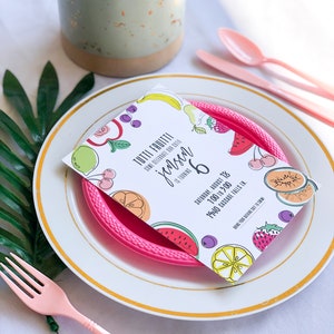 Puede incluir: Una tarjeta blanca con un dise&ntilde;o de frutas coloridas y el texto "Tutti Frutti" y "Jensen cumple 6 a&ntilde;os". La tarjeta est&aacute; sobre un plato de pl&aacute;stico rosa con un plato blanco debajo. El plato est&aacute; sobre un mantel blanco con una hoja verde en el fondo.